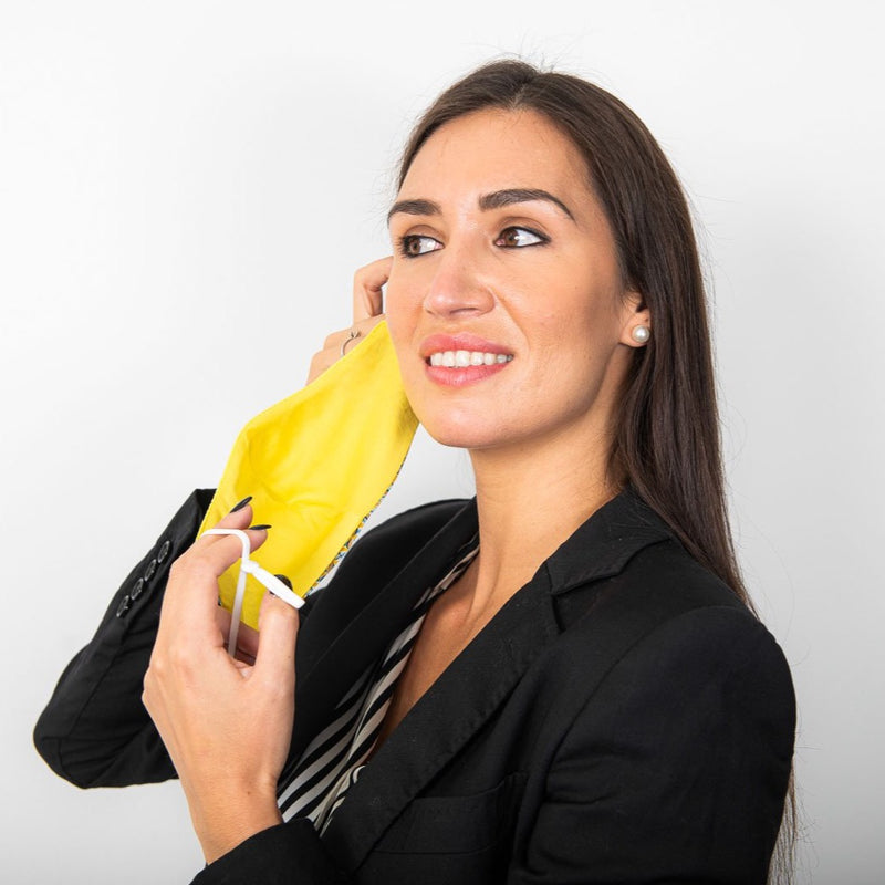 female model removing a mustard and blue geometric Liberty London Tana Lawn print face mask with a yellow lining and adjustable ear elastics.